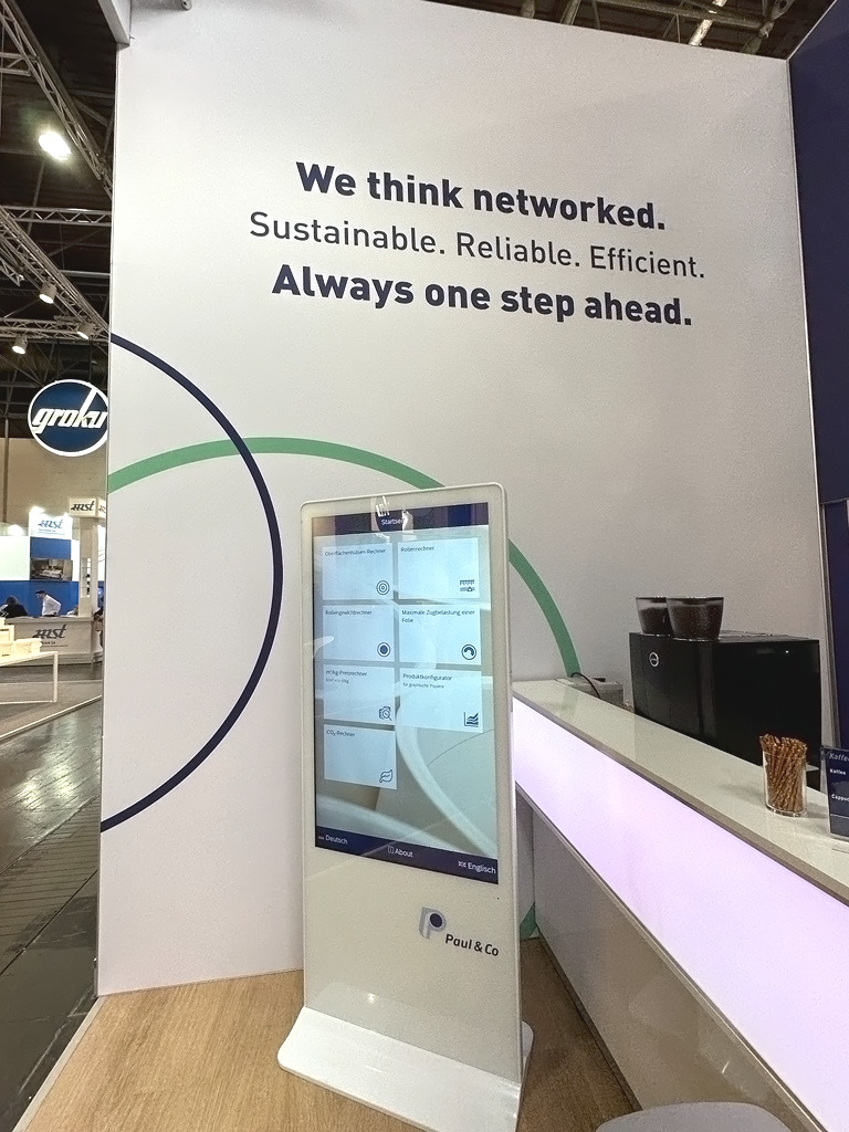 Paul & Co App auf großem Display am Messestand auf der Interpack 2023. Text auf der Wand im Hintergrund: "We think networked. Sustainable. Reliable. Efficient. Always one step ahead.